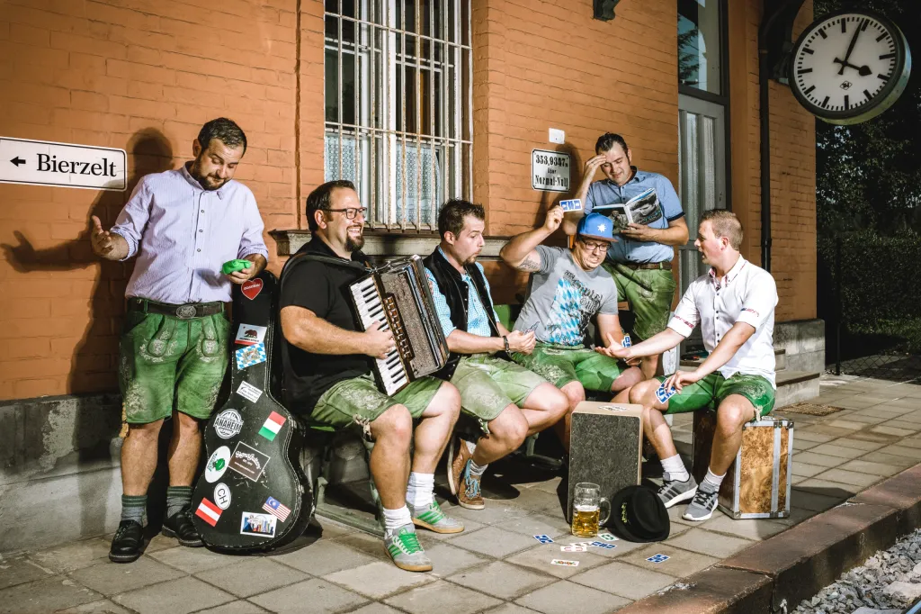 Fünf Musiker des Froschhaxn Express in Lederhosen vor einem Bahnhof, mit Akkordeon, Koffer und Wegweiser zum Bierzelt