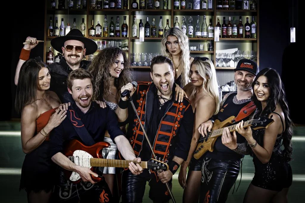 Acht Mitglieder der Partyband SKANDAL posieren vor einer Bar mit beleuchteter Flaschenwand, mit E-Gitarre und Mikrofon