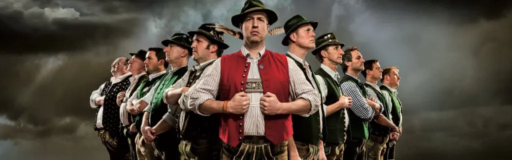 Kapelle Josef Menzl: Musiker in bayerischer Tracht mit Lederhosen und Trachtenhüten vor Wolkenhimmel