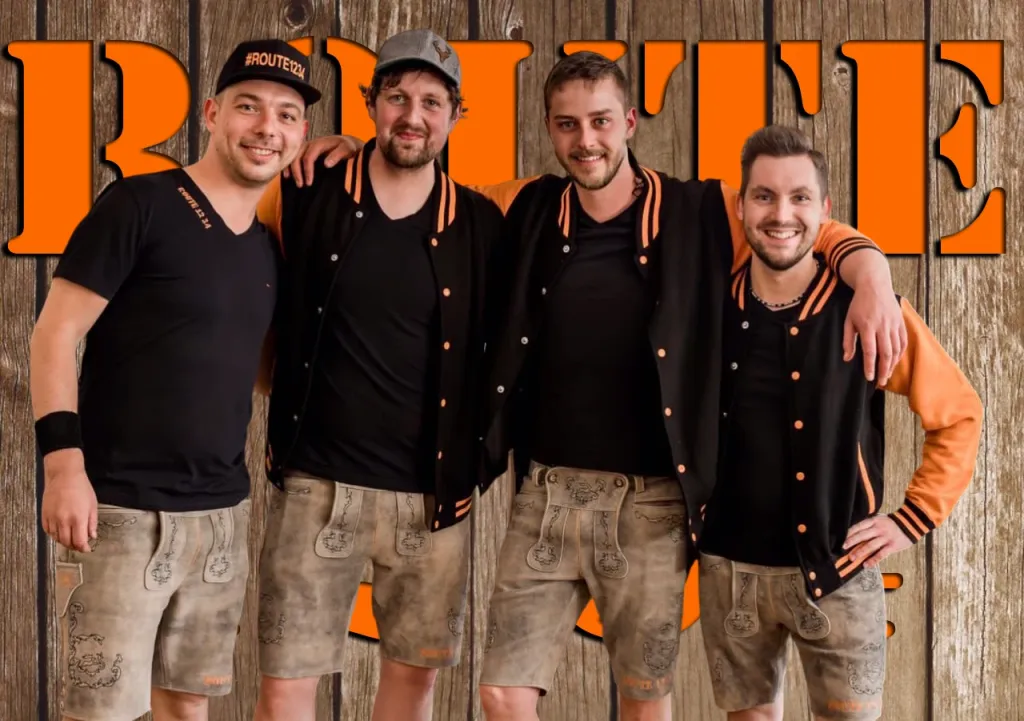 Vier Mitglieder der Band ROUTE 12 34 in schwarz-orangenen Pullovern und Lederhosen vor einer Holzwand mit „ROUTE“-Schriftzug