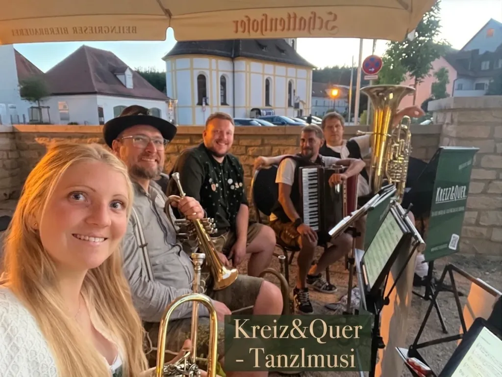 Fünf Musiker von Kreiz & Quer mit Trompete, Akkordeon und Tuba unter einem Sonnenschirm im Freien