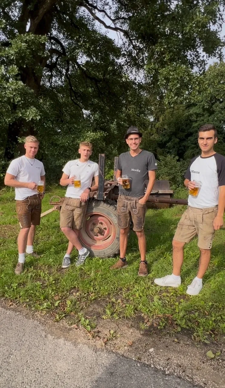 Vier junge Männer in Lederhosen, jeder mit einem Glas Bier, an einem alten Anhänger am Waldrand