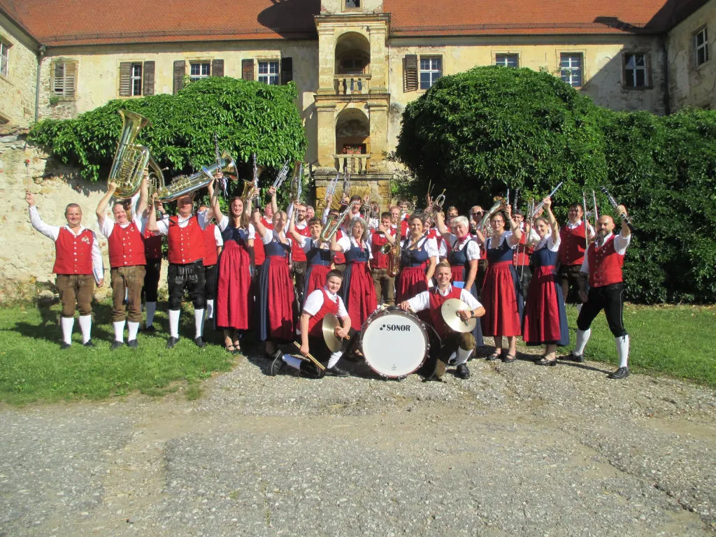 Jurablaskapelle Pilsheim in roten Trachtenwesten und Dirndln mit erhobenen Instrumenten vor einem historischen Gebäude