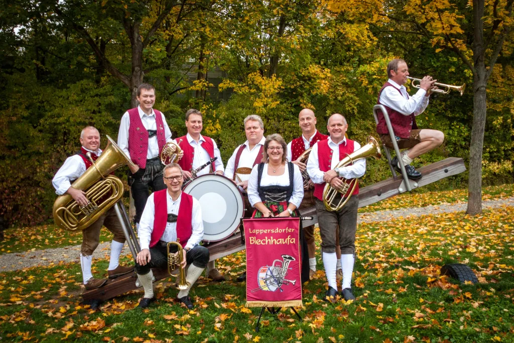 Neun Musiker der Lappersdorfer Blechhaufa in Tracht mit Blasinstrumenten auf einer Wippe im Herbstlaub