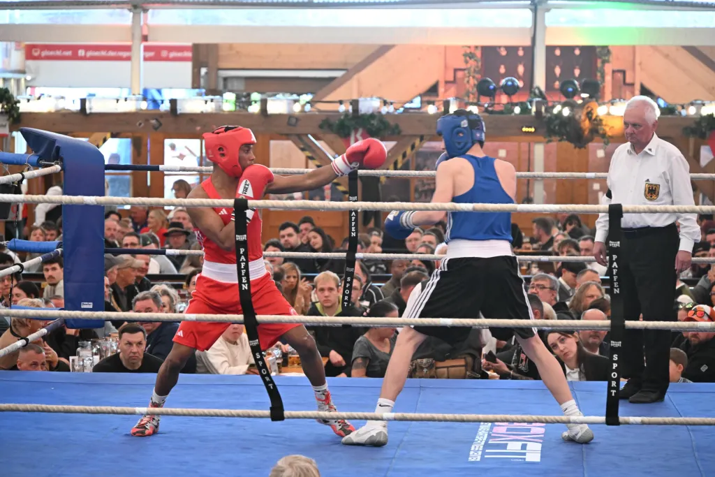 Zwei Boxer in roter und blauer Ausrüstung kämpfen im Ring eines vollbesetzten Festzelts, Kampfrichter am Rand