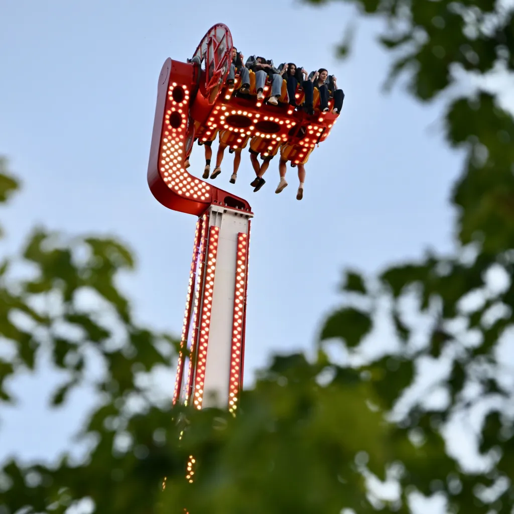Leuchtend roter Free-Fall-Tower mit Fahrgästen vor Abendhimmel zwischen Bäumen