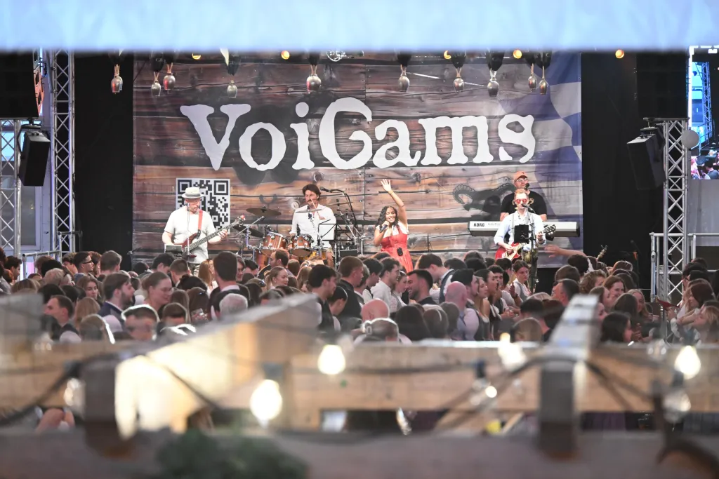 Die Band VoiGams spielt auf beleuchteter Bühne vor vollem Festzelt, Sängerin im roten Kleid winkt ins Publikum
