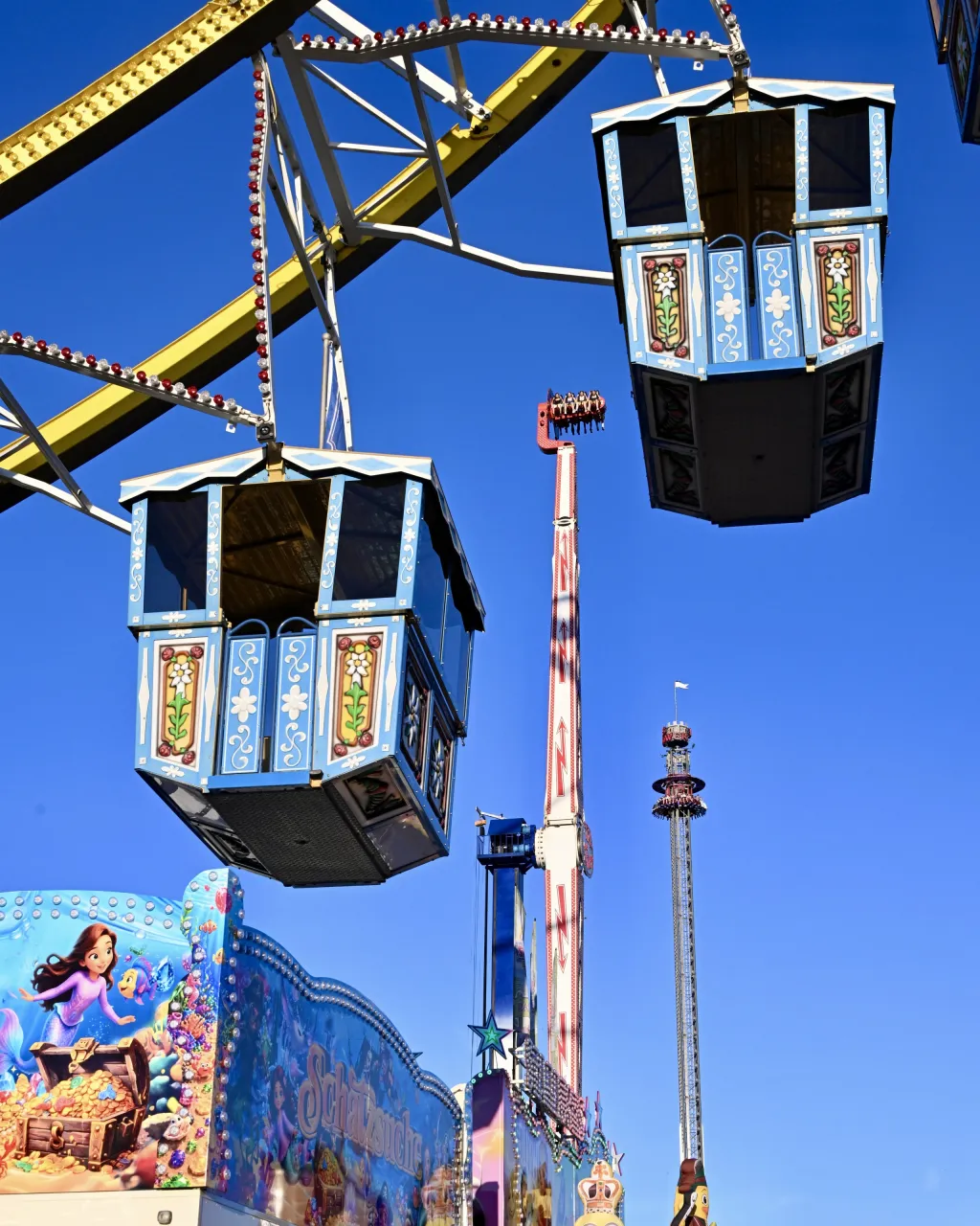 Zwei blaue Gondeln eines Riesenrads vor tiefblauem Himmel, dahinter ein hoher Freifallturm und Fahrgeschäfte