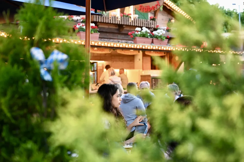 Blick durch grüne Büsche auf die beleuchtete Stüberl-Hütte mit blau-weißer Fahne und Gästen