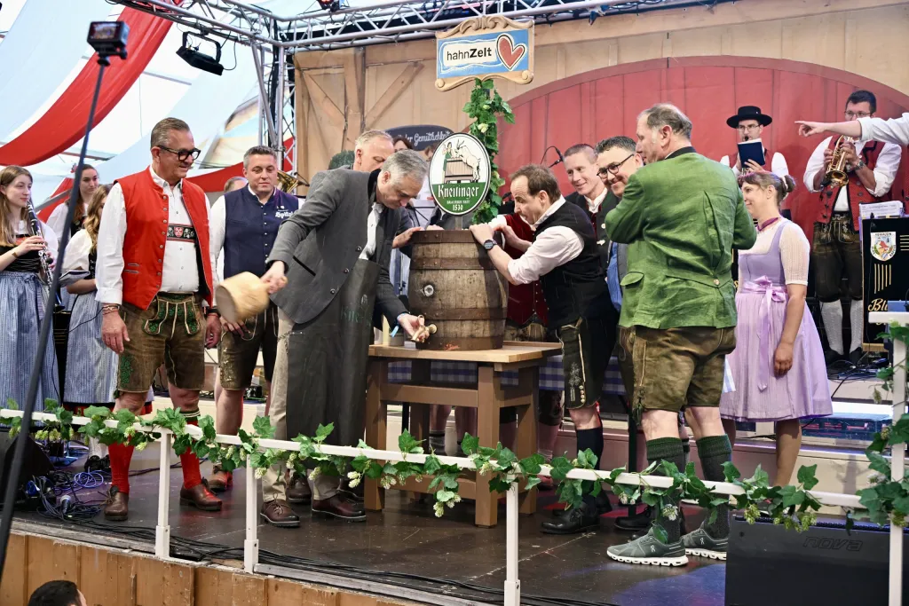 Fassanstich im Hahn Zelt: Mann schlägt den Zapfhahn ins Bierfass, umringt von Männern in Lederhosen