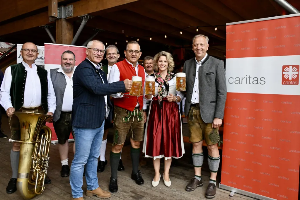 Gruppe in Tracht stößt mit Maßkrügen vor einem Caritas-Aufsteller im Festzelt an