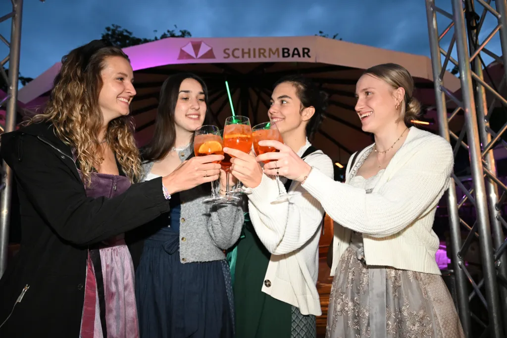 Vier junge Frauen in Dirndln stoßen mit Cocktailgläsern vor der Schirmbar an