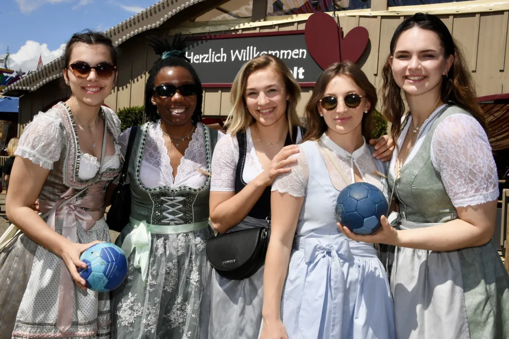 Fünf Frauen in hellen Dirndln posieren vor dem Eingang eines Festzelts mit Herz-Dekoration