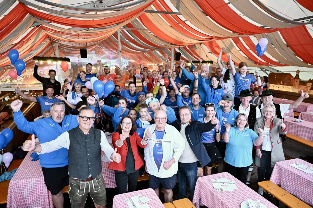 Große Gruppe Helfer in blauen T-Shirts und Tracht jubelt im festlich geschmückten Zelt