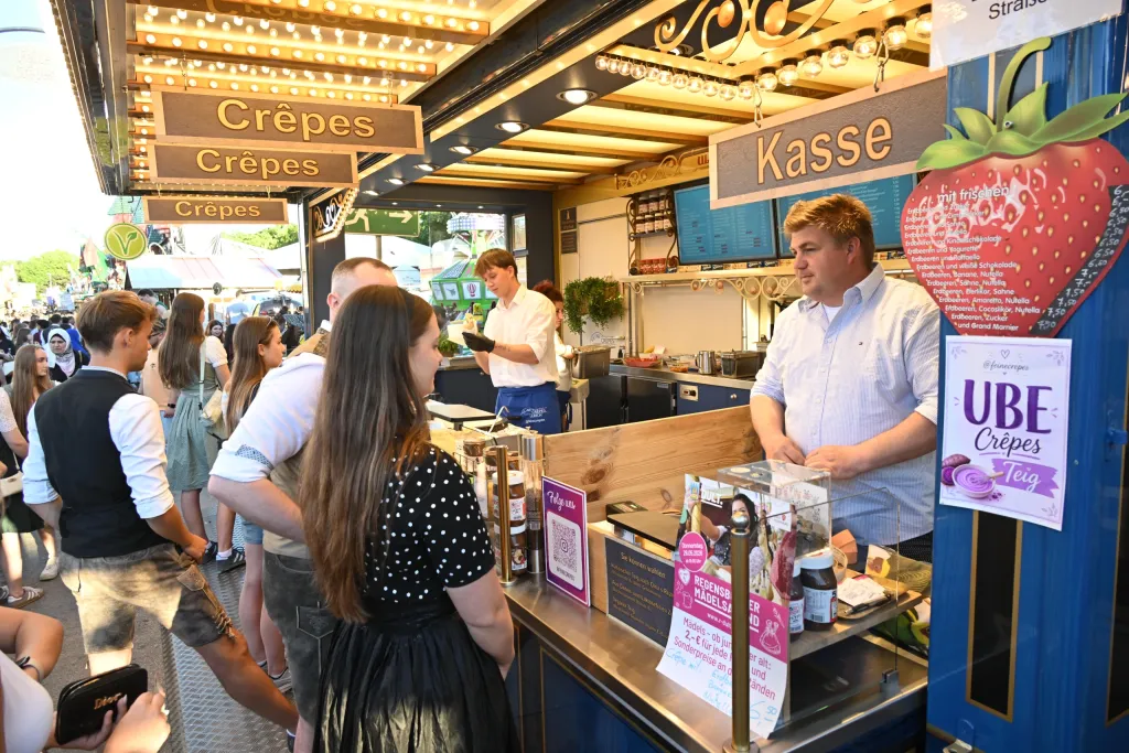 Junger Mann bedient die Kasse an einem bunt dekorierten Crêpes-Stand, eine Besucherin steht davor