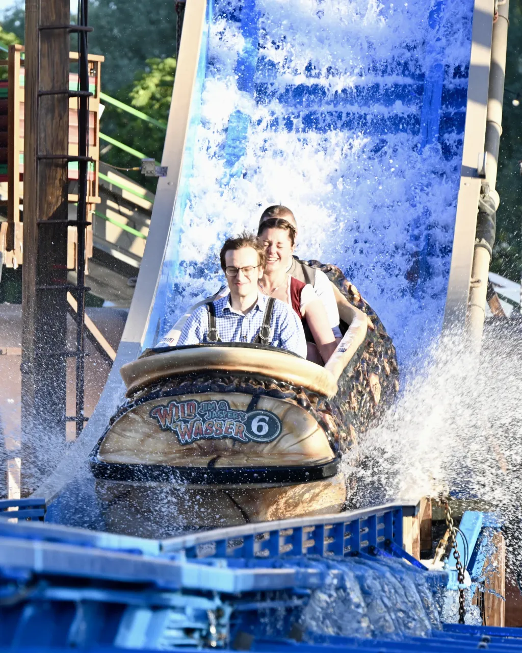 Zwei Besucher fahren lachend die Wasserrutsche des Fahrgeschäfts „Wildwasser“ hinunter, Fontänen spritzen