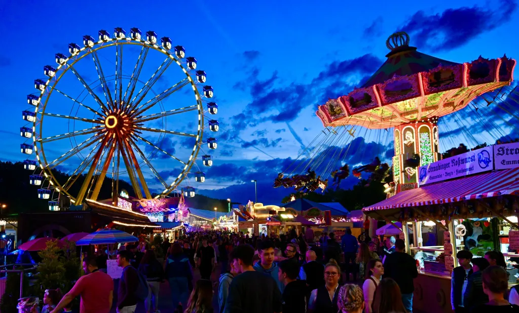 Beleuchtetes Riesenrad und Kettenkarussell auf der Dult bei Dämmerung mit Besuchermenge zwischen Buden