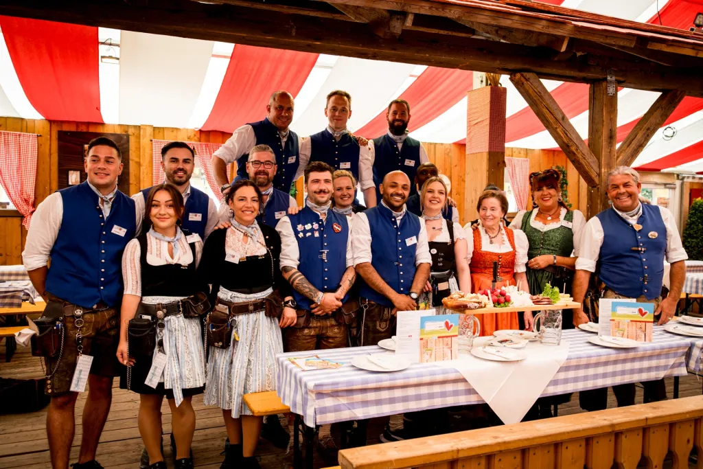 Service-Team in bayerischer Tracht mit blauen Westen und Dirndln im rot-weißen Hahn Zelt vor gedecktem Festzelttisch
