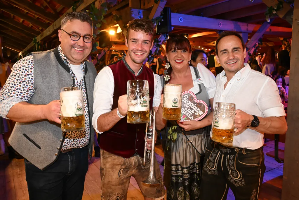 Zwei Männer in Tracht und eine Frau im Dirndl prosten lachend mit Maßkrügen im Festzelt