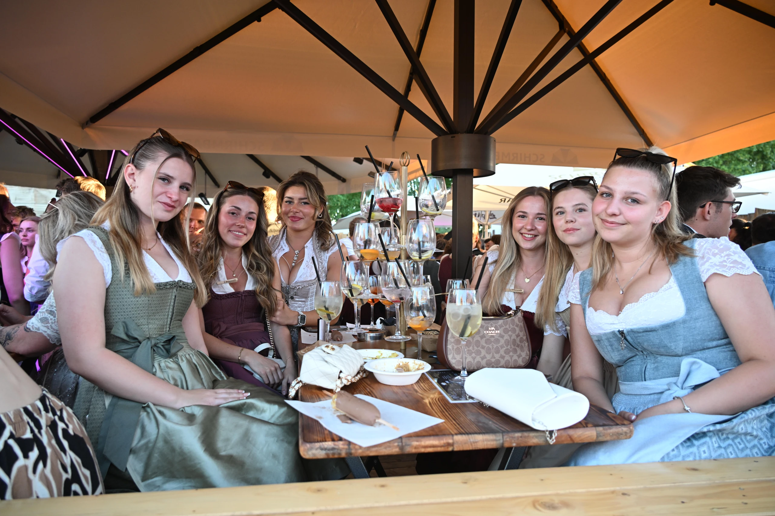 Sechs junge Frauen in Dirndln sitzen lächelnd unter Sonnenschirm an einem Biertisch mit Weinschorle-Gläsern