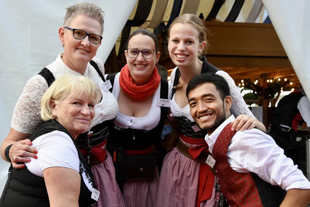 Festzeltbedienungen in Dirndl und Wirt umarmen sich lachend für Gruppenfoto