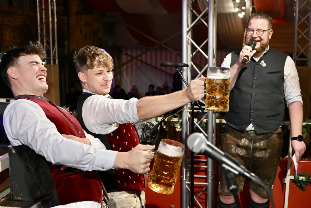 Zwei junge Männer in Tracht prosten Moderator mit Mikrofon und Maßkrug zu