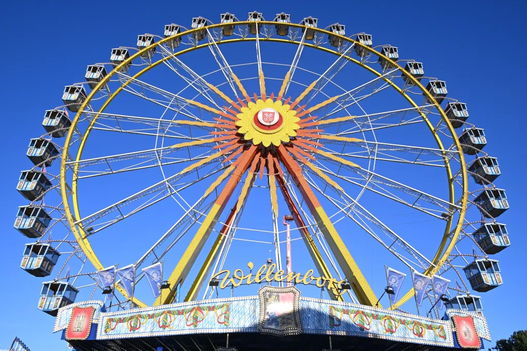 Großes gelb-oranges Riesenrad „Willenborg“ vor strahlend blauem Himmel, bayerische Wimpel am Eingang