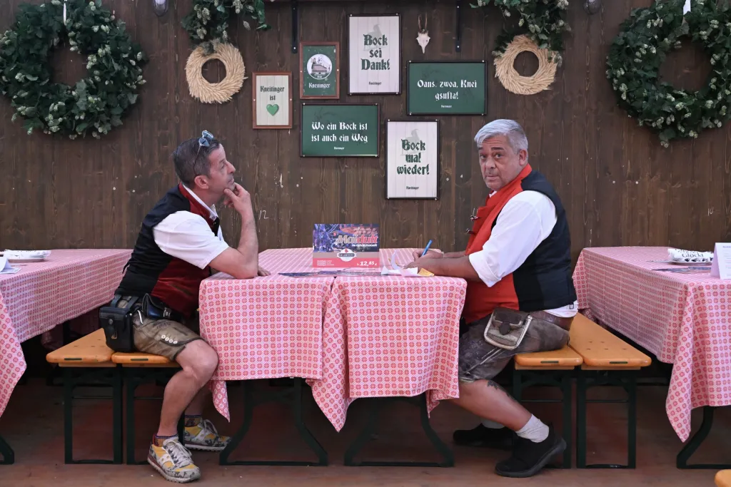 Zwei Männer in Tracht sitzen an Biertischgarnitur unter geschmückter Festzeltwand